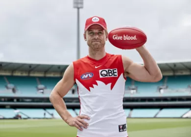 photo of sydney swans AFL player Dane Rampe in a stadium, he's wearing a Lifeblood cap and holding up a footy with the text 'Give blood' on it