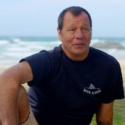 shark attack survivor Dave Pearson, talks to camera while sitting with the ocean behind him