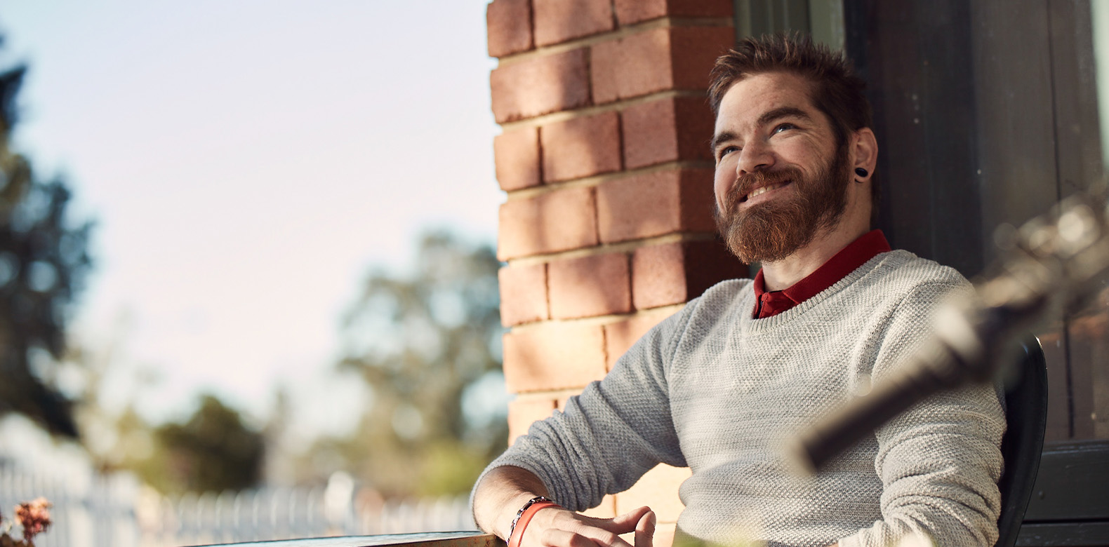 a man is sitting outside next to a wall and smiling, the afternoon sky can be seen in the background