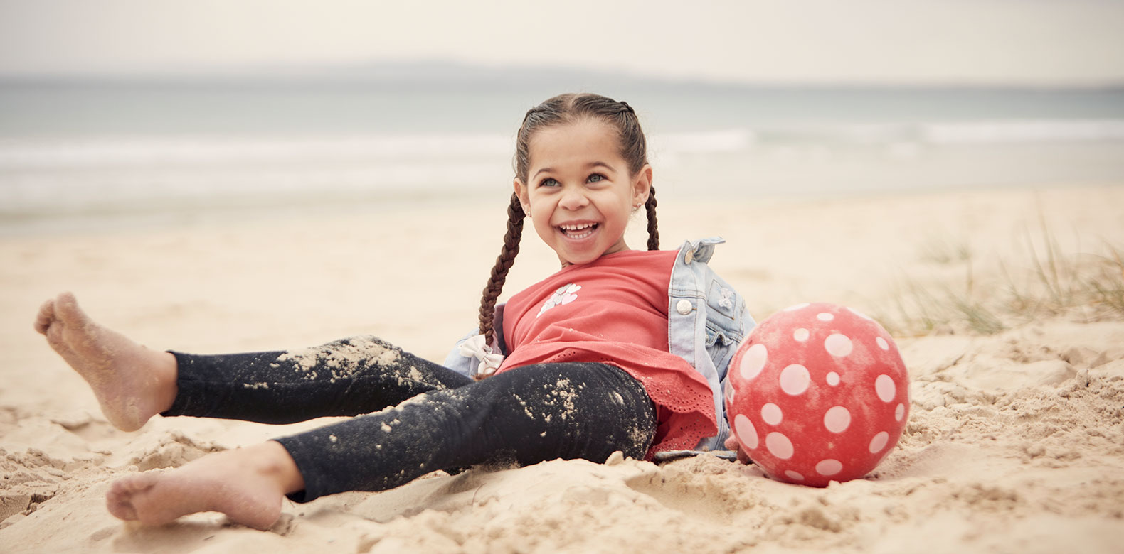 a girl playing with a beach ball on a beach sitting in the sand and smiling