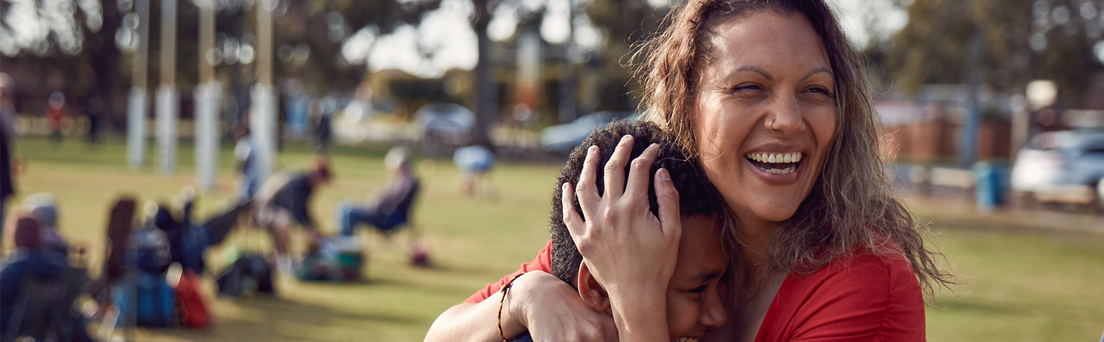 a mother is embracing her son and they are both laughing, a park can be seen in the background