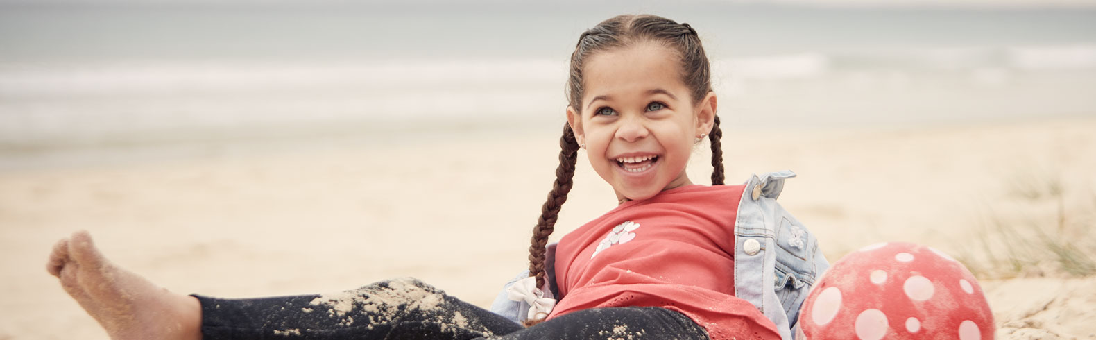a child sitting in the sand on a beach smiling, next to her is a red beach ball