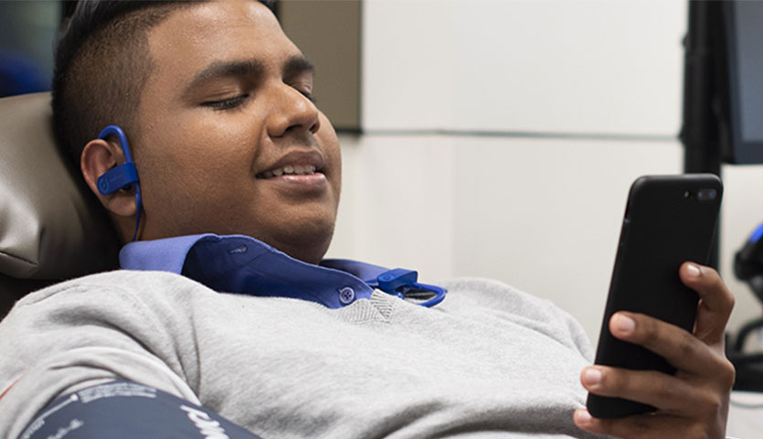 a donor sitting in a chair and donating, he is wearing earphones and looking at his phone smiling