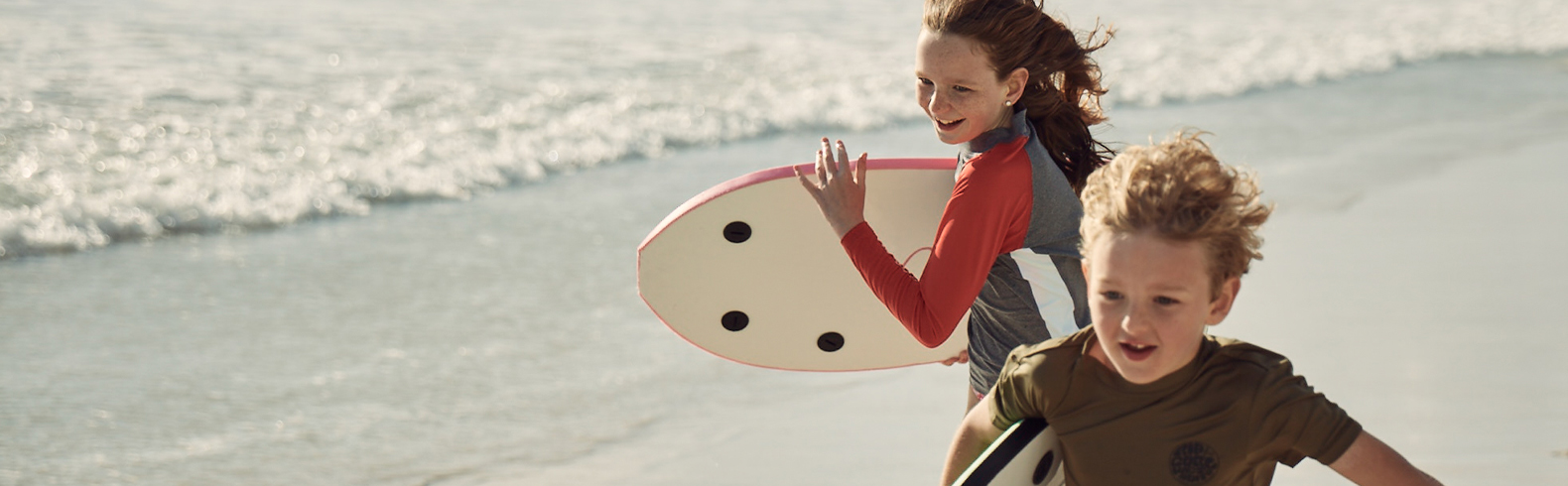 two kids smiling and running along the shore carrying bodyboards