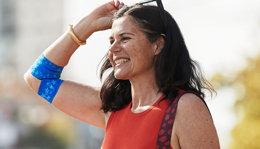 a woman is holding her sunglasses up and smiling off camera, on her elbow is a true colours designed bandage