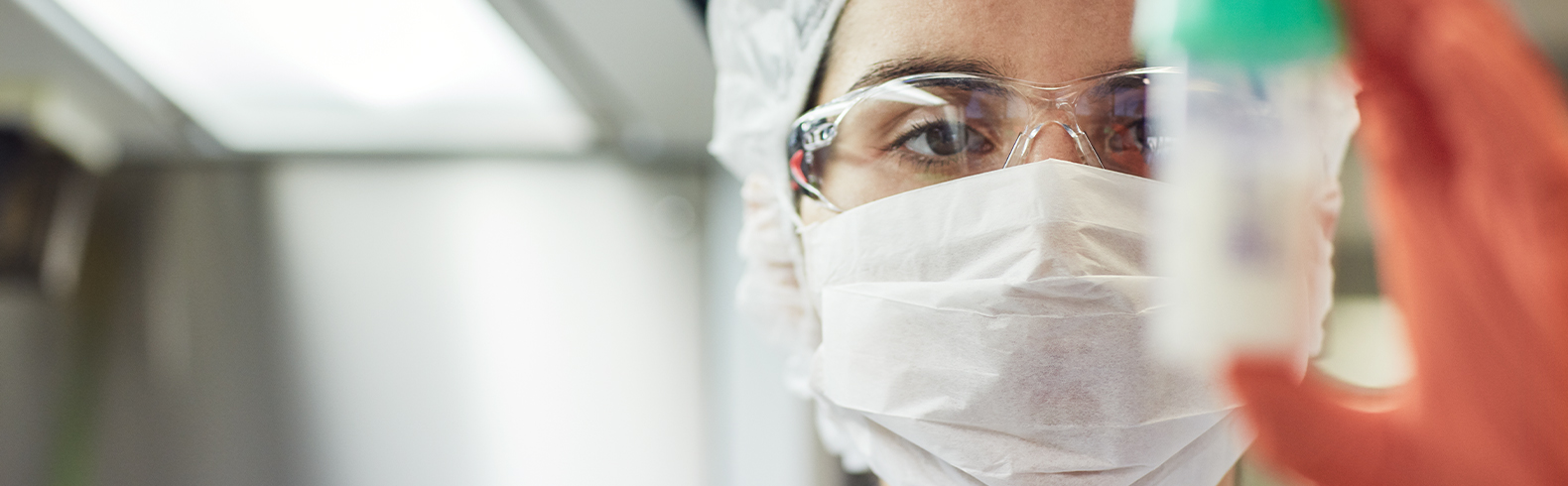 a scientist is wearing a facemask and a hairnet and is holding up a milk sample jar