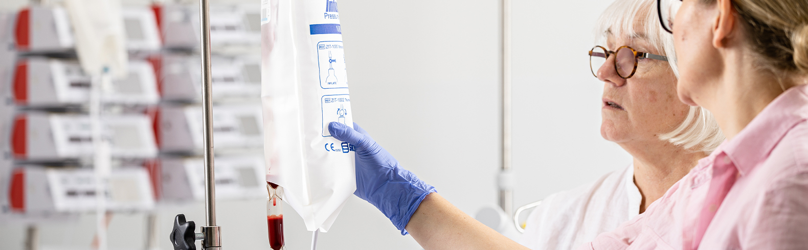 two nurses wearing pink gowns and blue gloves are examining an IV and discussing it