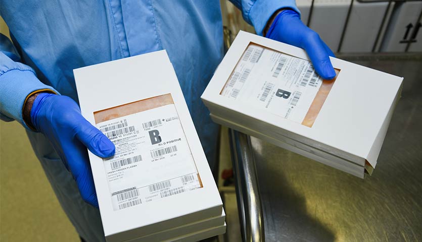 a lab worker is wearing a blue gown and gloves and is holding two boxes of blood packs marked B positive