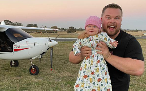 man holding a baby smiling at camera in front of an airplane