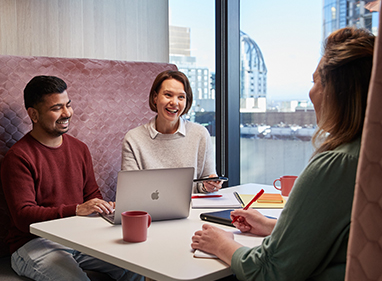 three work colleagues are sitting in a booth and smiling mid conversation, a laptop and notepads are on the table