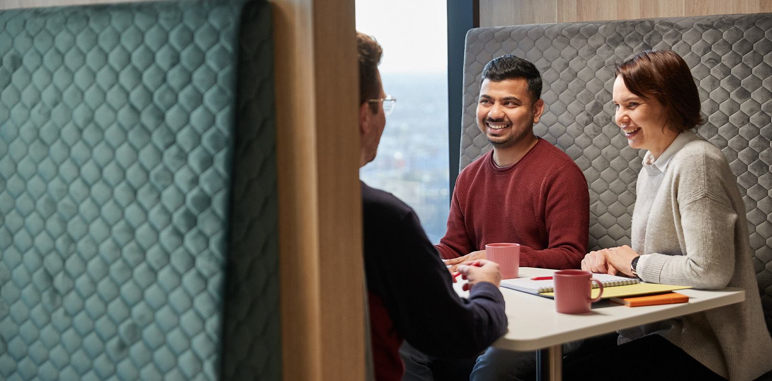 three work colleagues are sitting in a booth and smiling mid conversation, a laptop and notepads are on the table