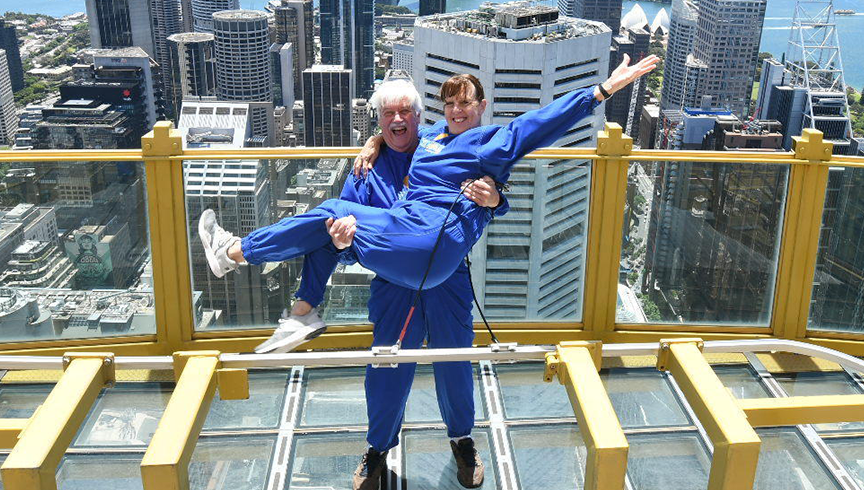Man holding up woman wearing blue overalls at the top of a building