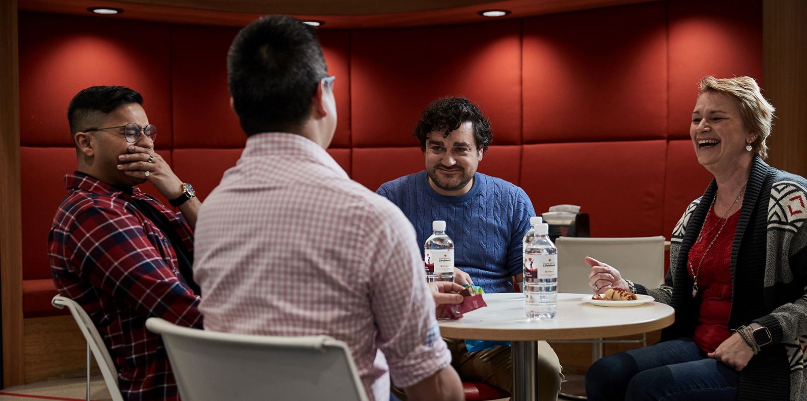 four people are seated at a table smiling and laughing, on table there is water bottles, crackers and a sausage roll with sauce