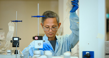 scientist is working on some equipment and holding up a donation bag, they are wearing a blue gown and gloves