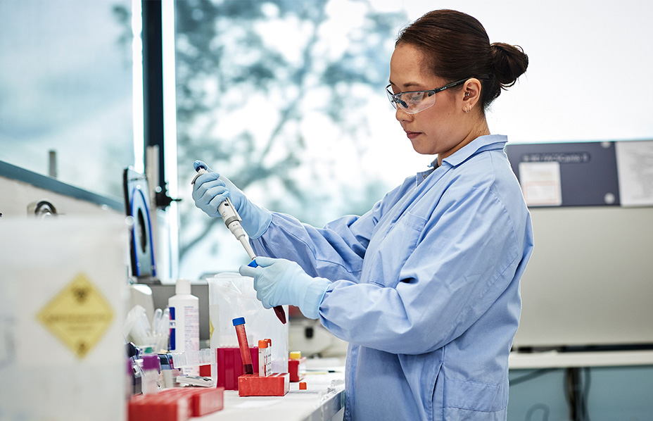 photograph of scientist wearing blue lab coat safety glasses and gloves holding a pipette