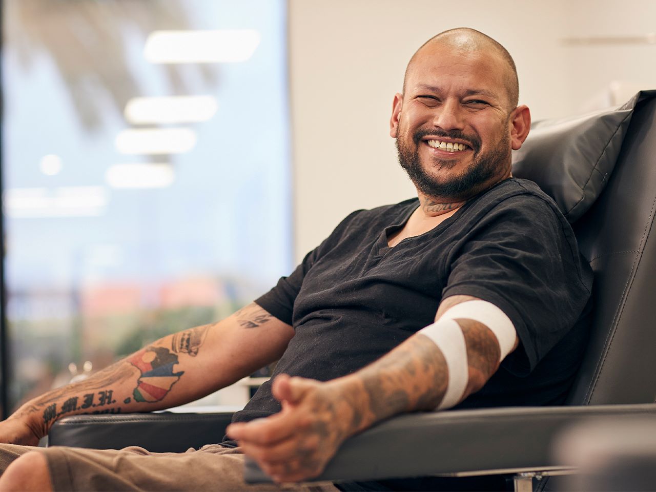 a donor is seated in a chair after a donation with a bandage around his elbow and smiling at someone off camera