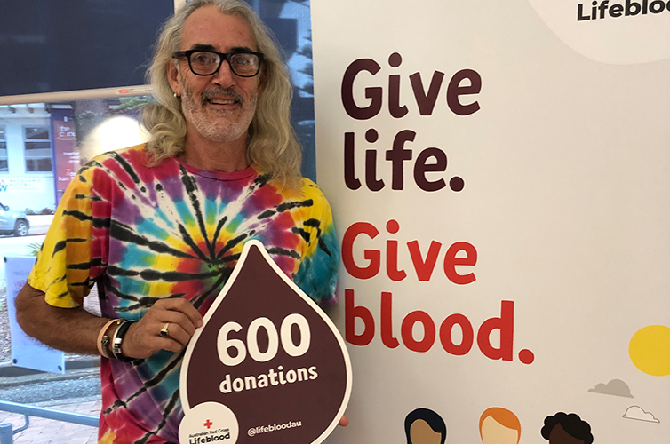 Donor Bruce smiling and holding a sign with 600 donations written on it, he stands next to a banner with give life give blood written on it