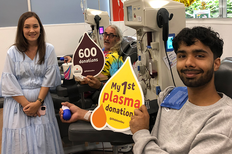 Taylor standing next to Bruce and Swatip while they're donating, bruce holds a sign with 600 donations written on it and swatip is holding a sign with my 1st plasma donation on it