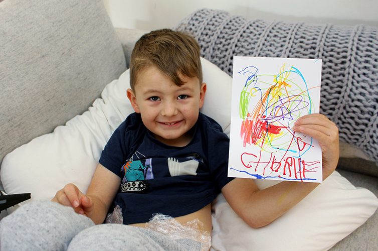 boy sitting on grey couch smiling and holding up his drawing