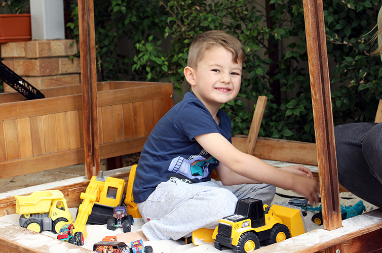 Smiling boy in sand pit, playing with toy trucks 