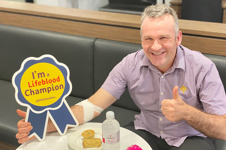 James is sitting at a table smiling and giving a thumbs up, he is holding a sign saying I am a Lifeblood champion