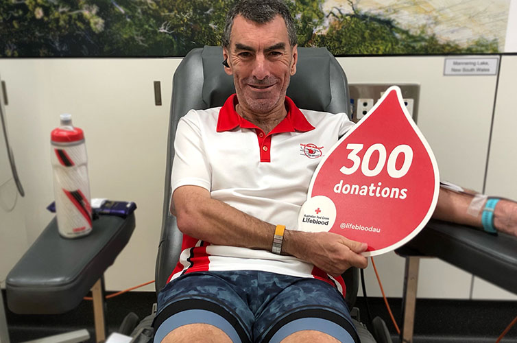 donor Jarrod is smiling in the donor chair holding up a 300 donations card shaped like a red blood drop