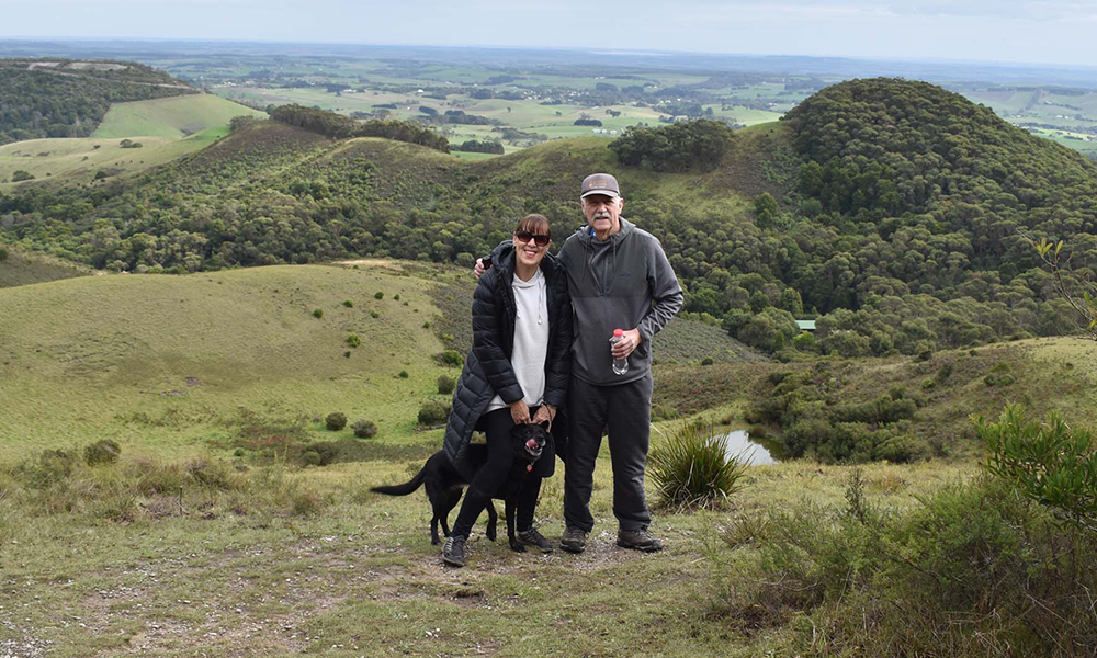 man, woman and black dog smiling at camera with a green landscape in the background