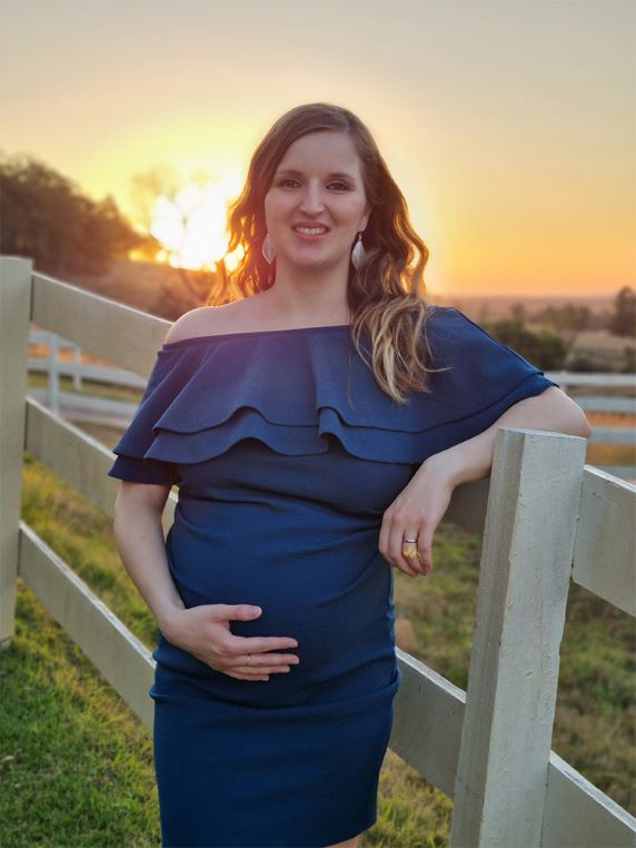 donor katarina is smiling at the camera and leaning her arm on a fence, in the background is a natural landscape and yellow sunset, she is wearing a blue dress