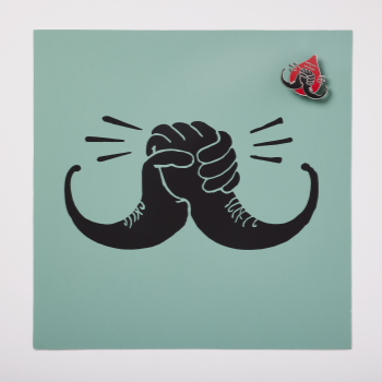 photo of the travis garone design, it's a stylised illustration of two hands holding one another in a moustache shape on a blue background, on top of the design is a pin in a blood drop shape featuring the design