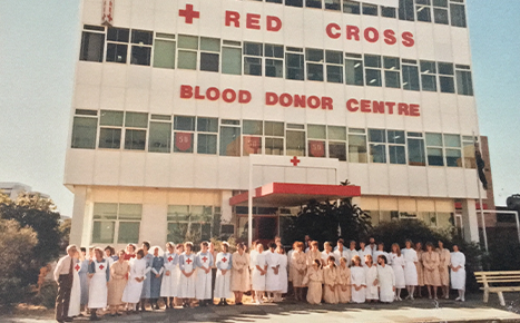 1980s colour photo of a large group of women, some of who are nurses, and a man in a tie standing outside a tall building saying "Red Cross Blood Donor Centre" 