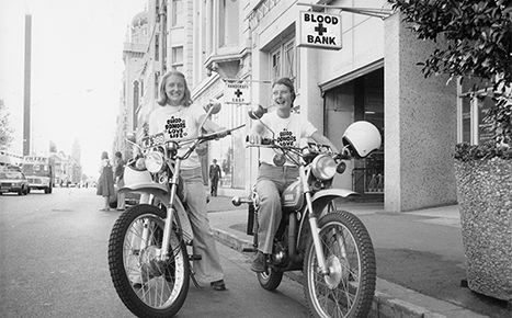 1970s black and white image. Two women on motorcycles wearing t-shirts saying "Blood Donors Love Life"
