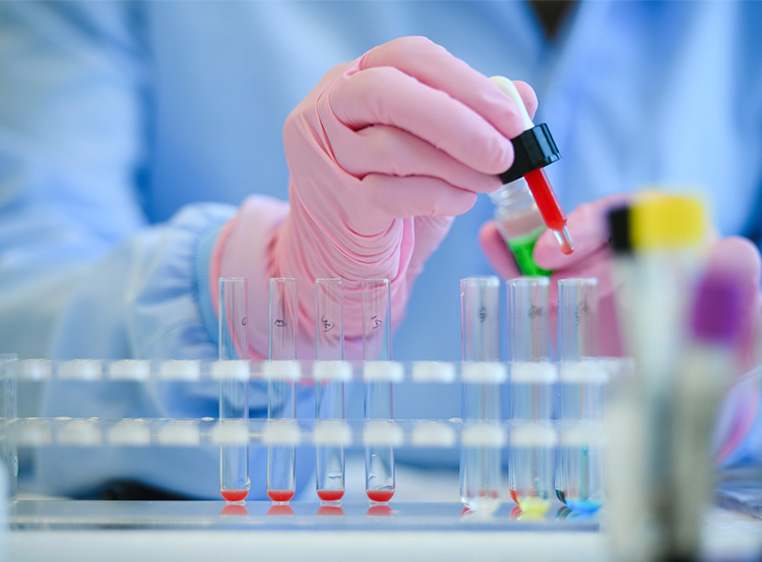 photo of a scientist wearing a blue gown and pink gloves holding a dropper over a row of test tubes