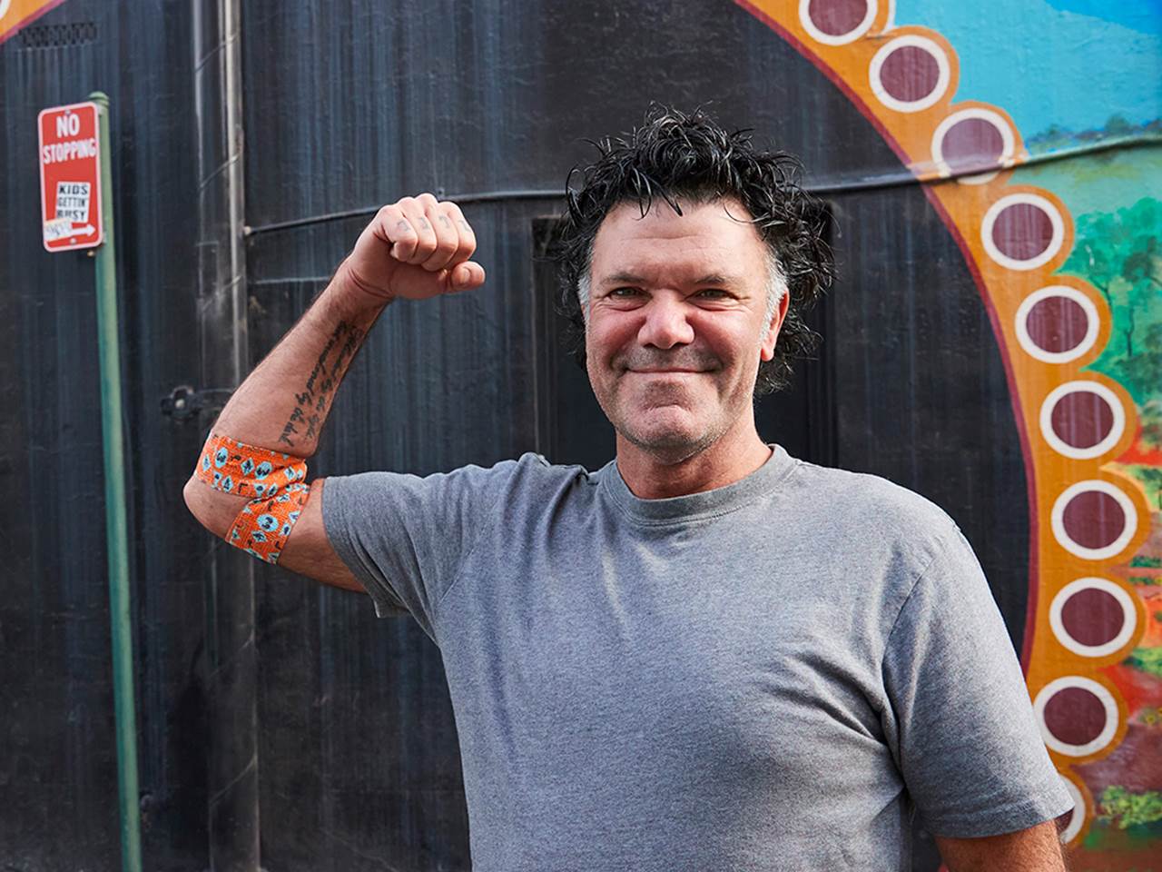 a photo of artist blak douglas smiling and holding his right arm up in a flex pose, around his elbow is the bandage he designed