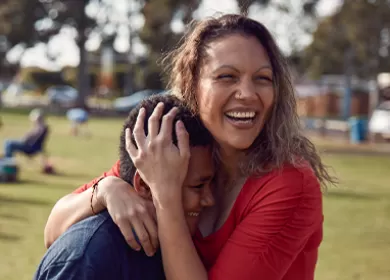 a mother is embracing her son and they are both laughing, a park can be seen in the background