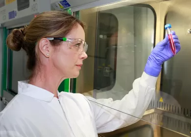 a scientist is wearing a white coat and purple gloves, she is looking at a vial of blood she's holding up 