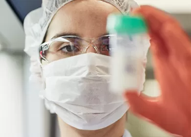 a scientist is wearing a facemask and a hairnet and is holding up a milk sample jar