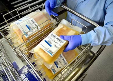 a lab worker wearing a blue gown and gloves holding bags of plasma in a trolley