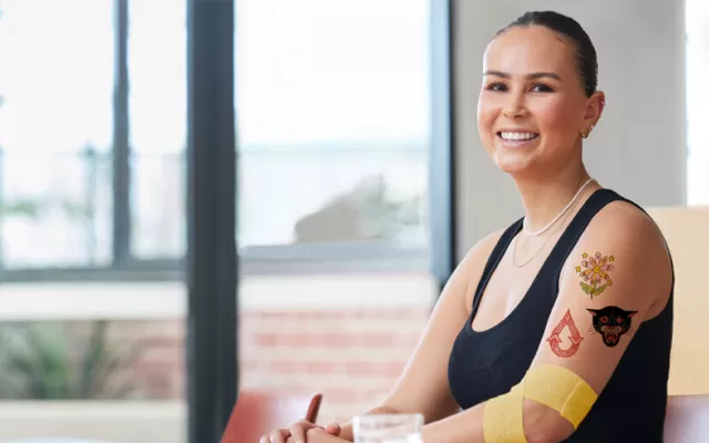 photograph of a woman sitting at a table and smiling, she has three temporary tattoos on her left arm and a yellow bandage around her elbow