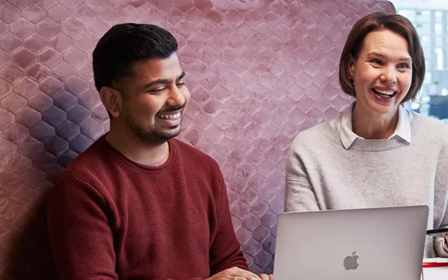 three work colleagues are sitting in a booth and smiling mid conversation, a laptop and notepads are on the table