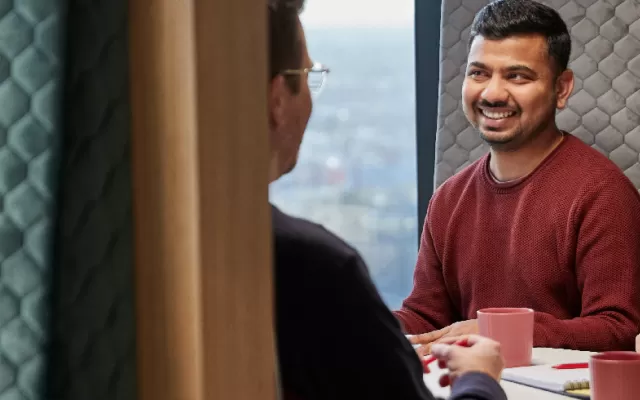 three work colleagues are sitting in a booth and smiling mid conversation, a laptop and notepads are on the table