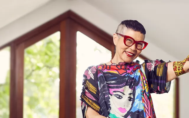 photo of Jenny Kee inside a room which looks out onto trees, Jenny is wearing a colourful shirt and red glasses and smiling, her left arm is raised and around her elbow is the bandage with her design on it
