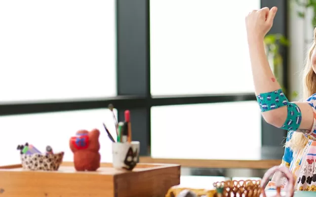 designer min pin is seated at a table wearing a colourful top and smiling, her left arm is in a flex pose and around her elbow is the bandage she designed