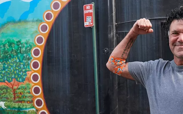 a photo of artist blak douglas smiling and holding his right arm up in a flex pose, around his elbow is the bandage he designed