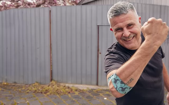 photo of Travis Garone smiling at the camera and holding up his right arm, around his elbow is the bandage he designed, he's wearing a black t-shirt and standing in a laneway