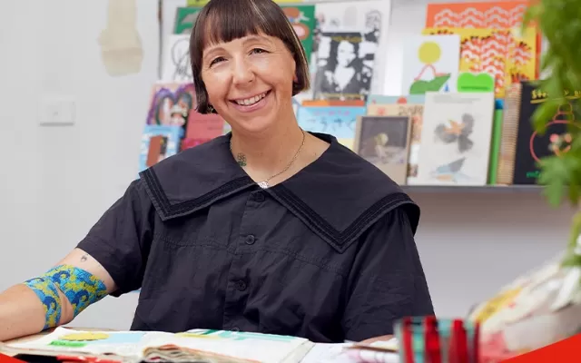photo of artist Beci Orpin seated at a desk and smiling at the camera, she is wearing a black blouse and around her right elbow is the bandage she designed
