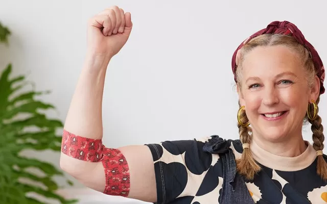 photo of artist Madeleine Stamer smiling at the camera with her right hand raised, around her elbow is the bandage with her design