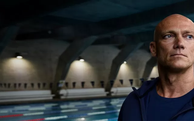 photograph of swimmer michael klim wearing a blue t shirt and hoodie, in the background behind him is an indoor swimming pool