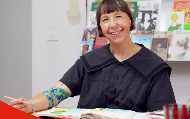 photo of artist Beci Orpin seated at a desk and smiling at the camera, she is wearing a black blouse and around her right elbow is the bandage she designed