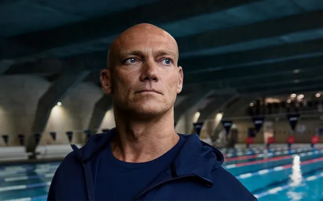 Michael Klim looking off camera and wearing a blue t shirt and hoodie, in the background is a an indoor swimming pool