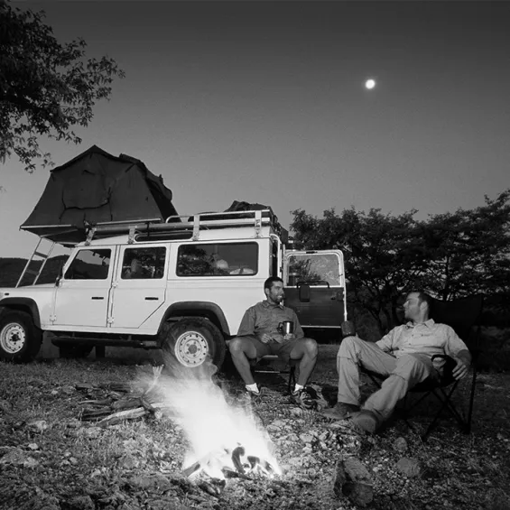 two men sitting and talking by a campfire with a four wheel drive parked behind them